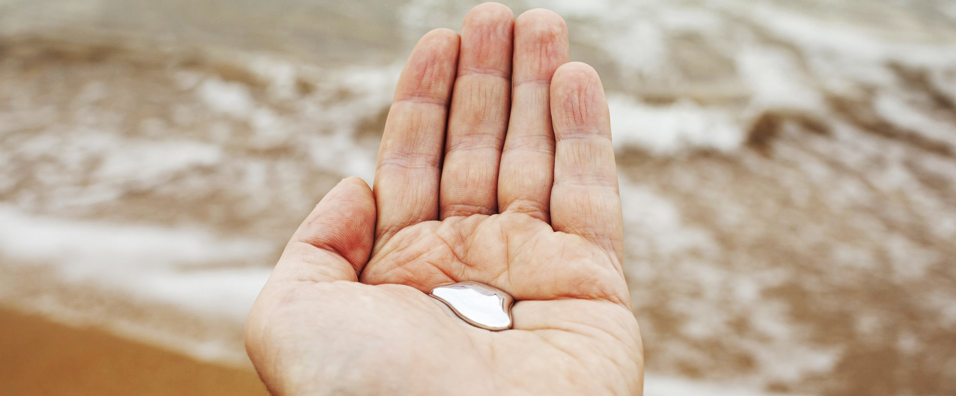 A hand holding a drop of liquid mercury with ocean water in the background